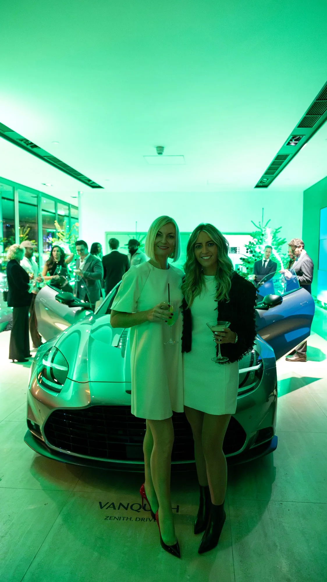 Guests beside the new Aston Martin Vanquish at the Mayfair launch, drinks in hand in the green lit showroom