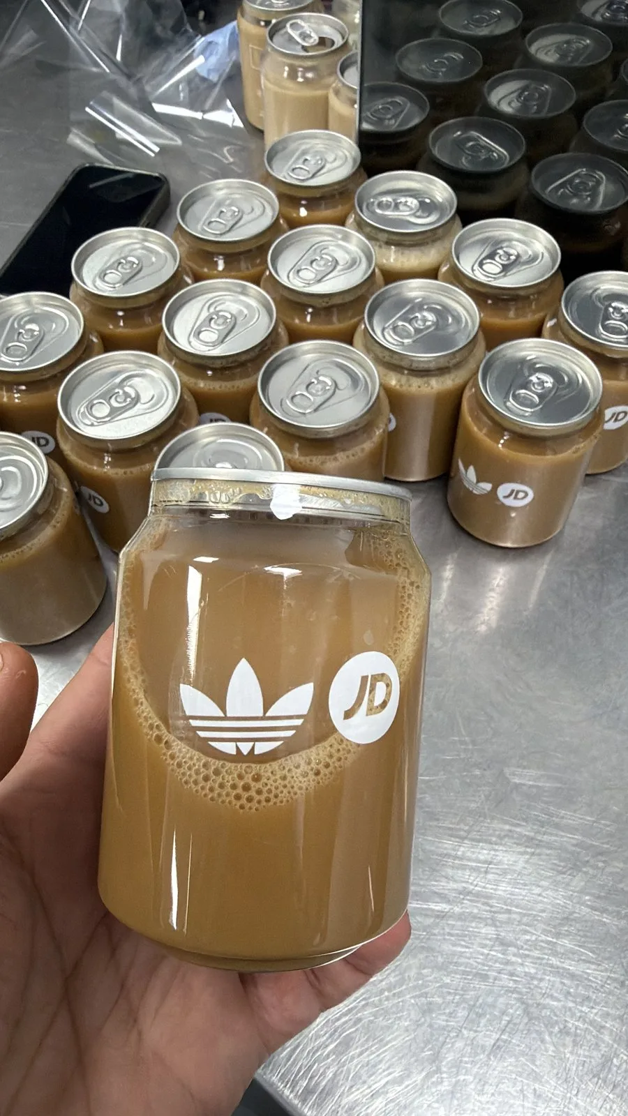 A branded iced latte can carrying the Adidas trefoil and JD Sports logo, held over rows of finished cans on the production table