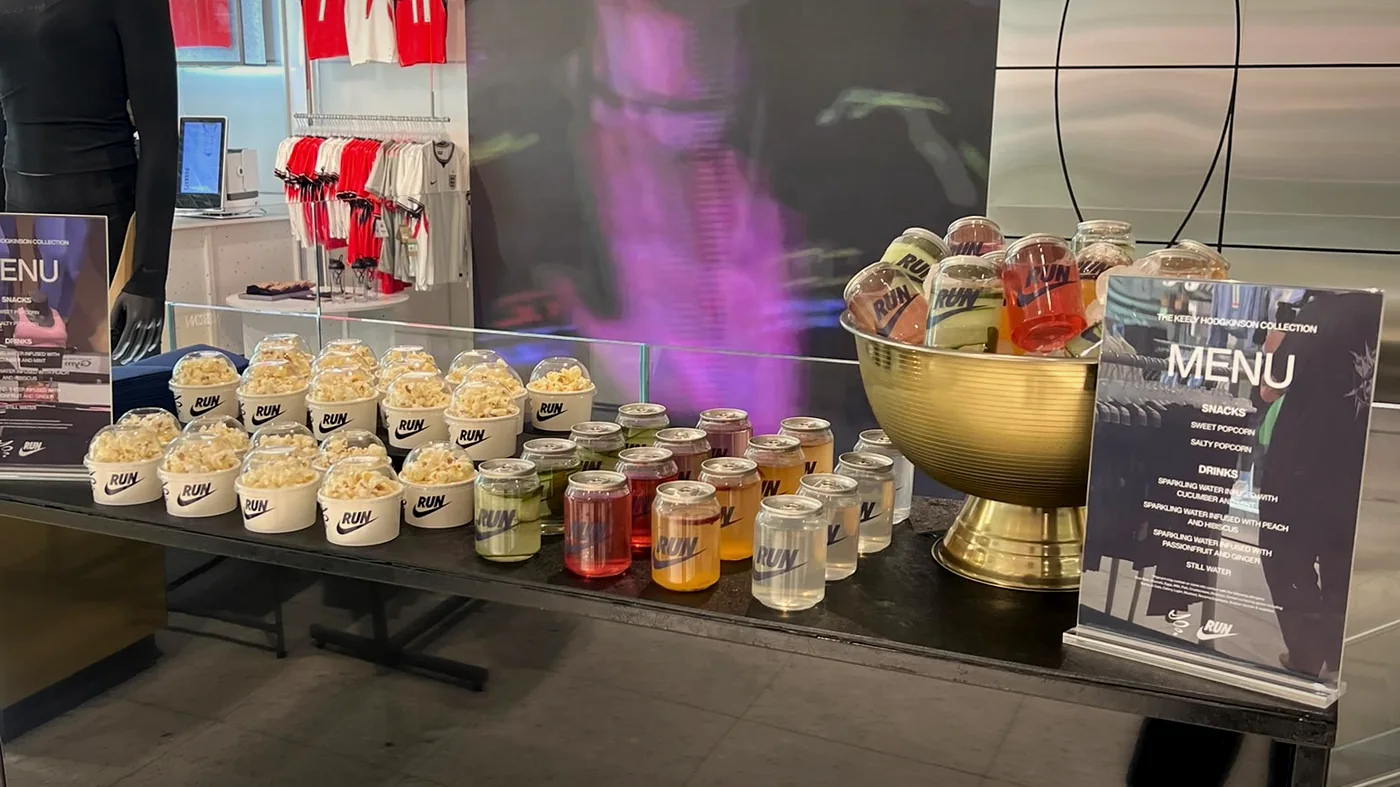 A Nike store activation table set with branded popcorn tubs, a gold ice bowl and rows of clear RUN cans beside a printed menu card.