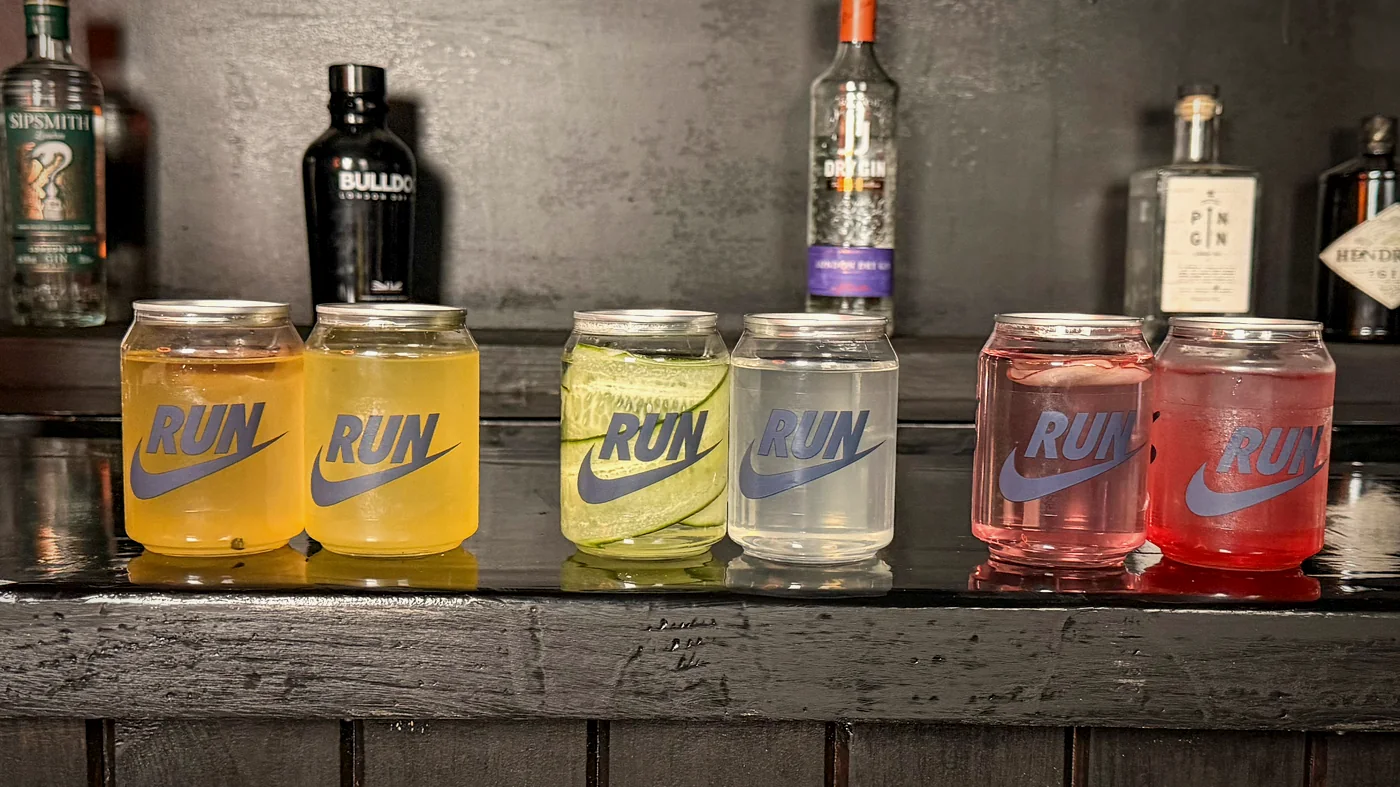 Six clear Mr Flavour cans in a row on a bar, each printed with the Nike RUN mark, filled with differently coloured infused waters.
