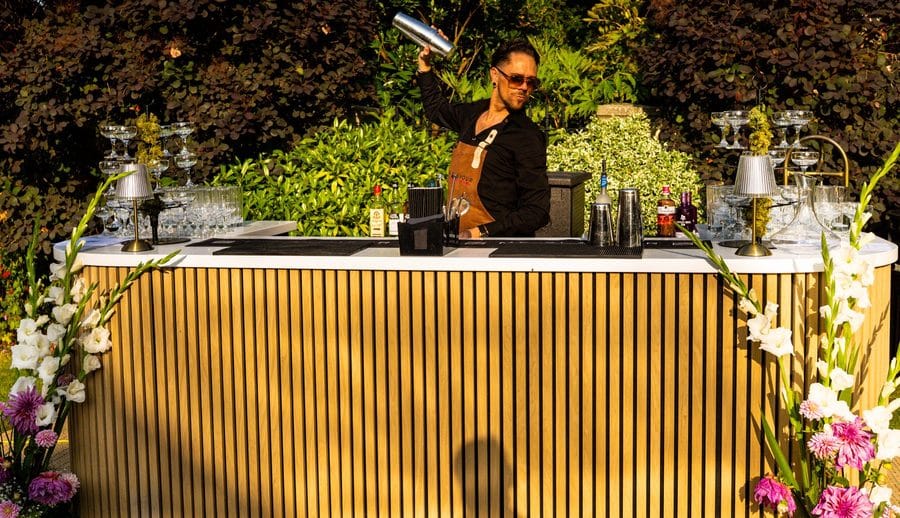 John shaking a drink behind a gold slatted bar dressed with flowers